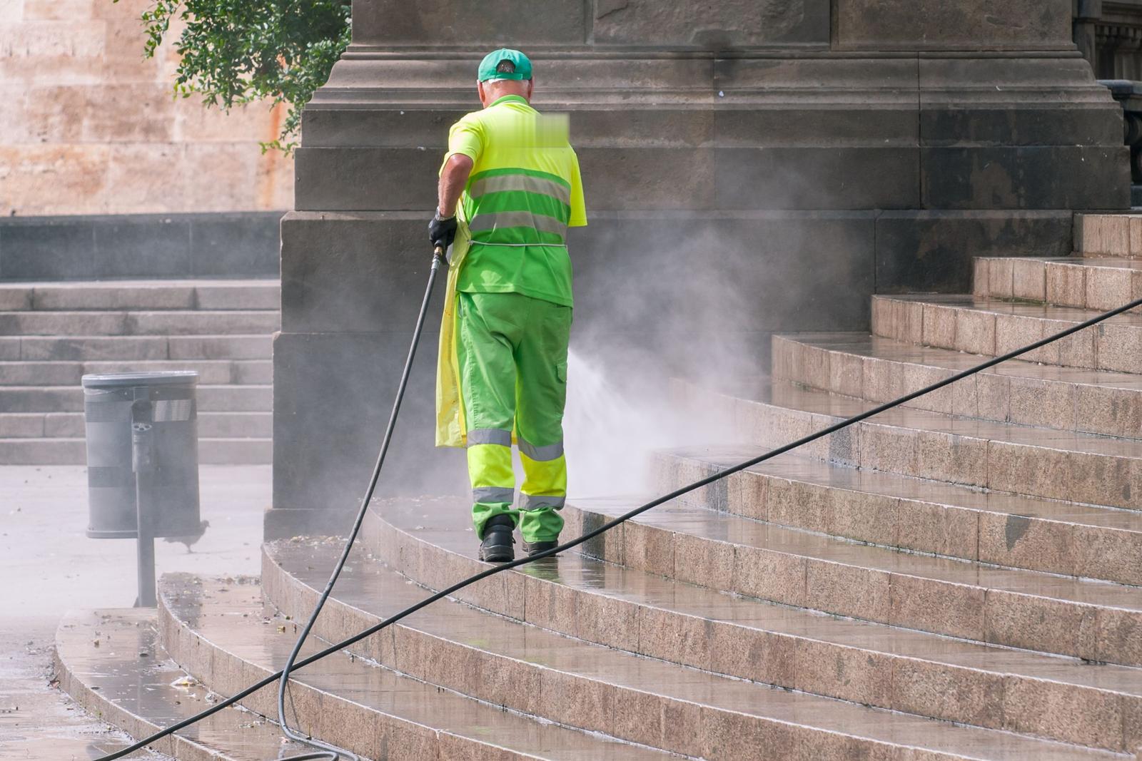 Worker seen from behind pressure washing wide stone steps, spray mist rising off the stone