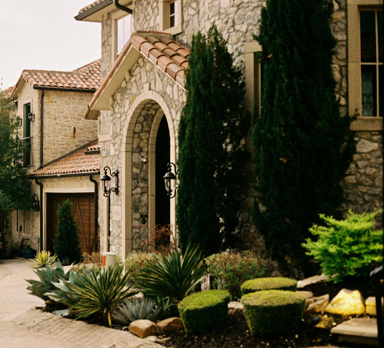 Stone-clad custom home in Dallas with a tall arched stone entry, tile roof and drought-tolerant landscaping