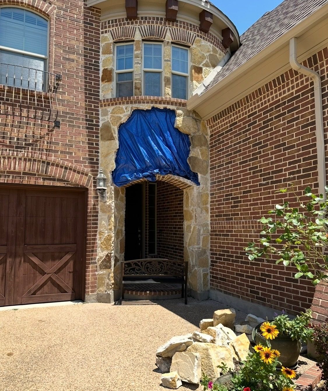 Brick house with a stone tower; the arch over the front entry is covered with a blue tarp during the repair, with loose stone on the drive