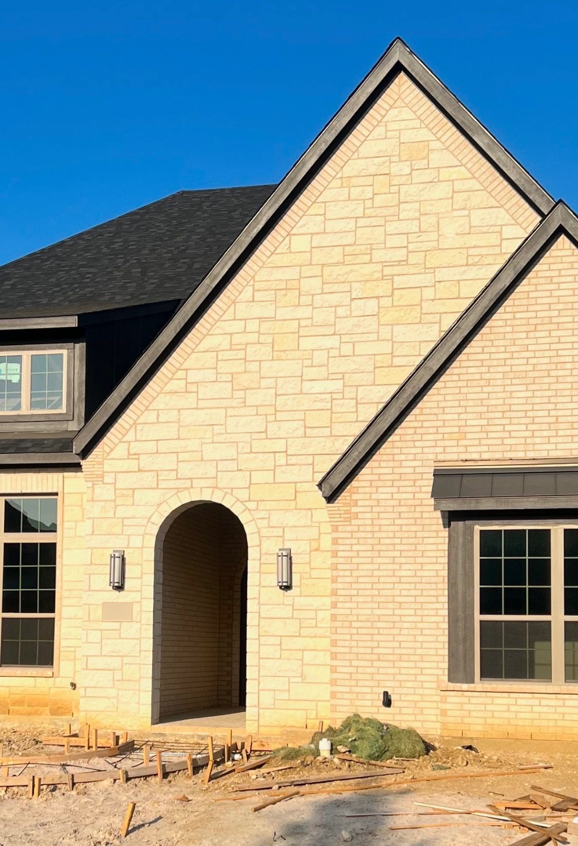Gable of a new home in cream stone with brick corners and an arched front entry under a blue sky