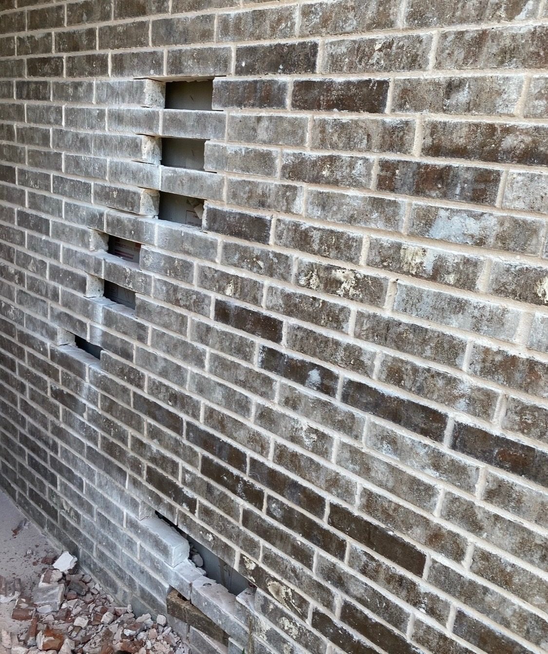 Gray brick wall with several damaged bricks cut out near the base, before the repair